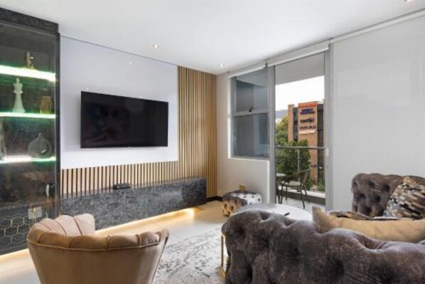 Ein modernes Wohnzimmer mit einem flachbildfernseher an der Wand, einer Wand aus Naturstein, einem großen Fenster und einer gemütlichen Sitzecke mit Sofa und Hundebett.