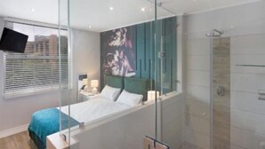 A modern hotel room with a glass shower enclosure, a bed with a blue bedspread, a mounted TV, and a large window with blinds.