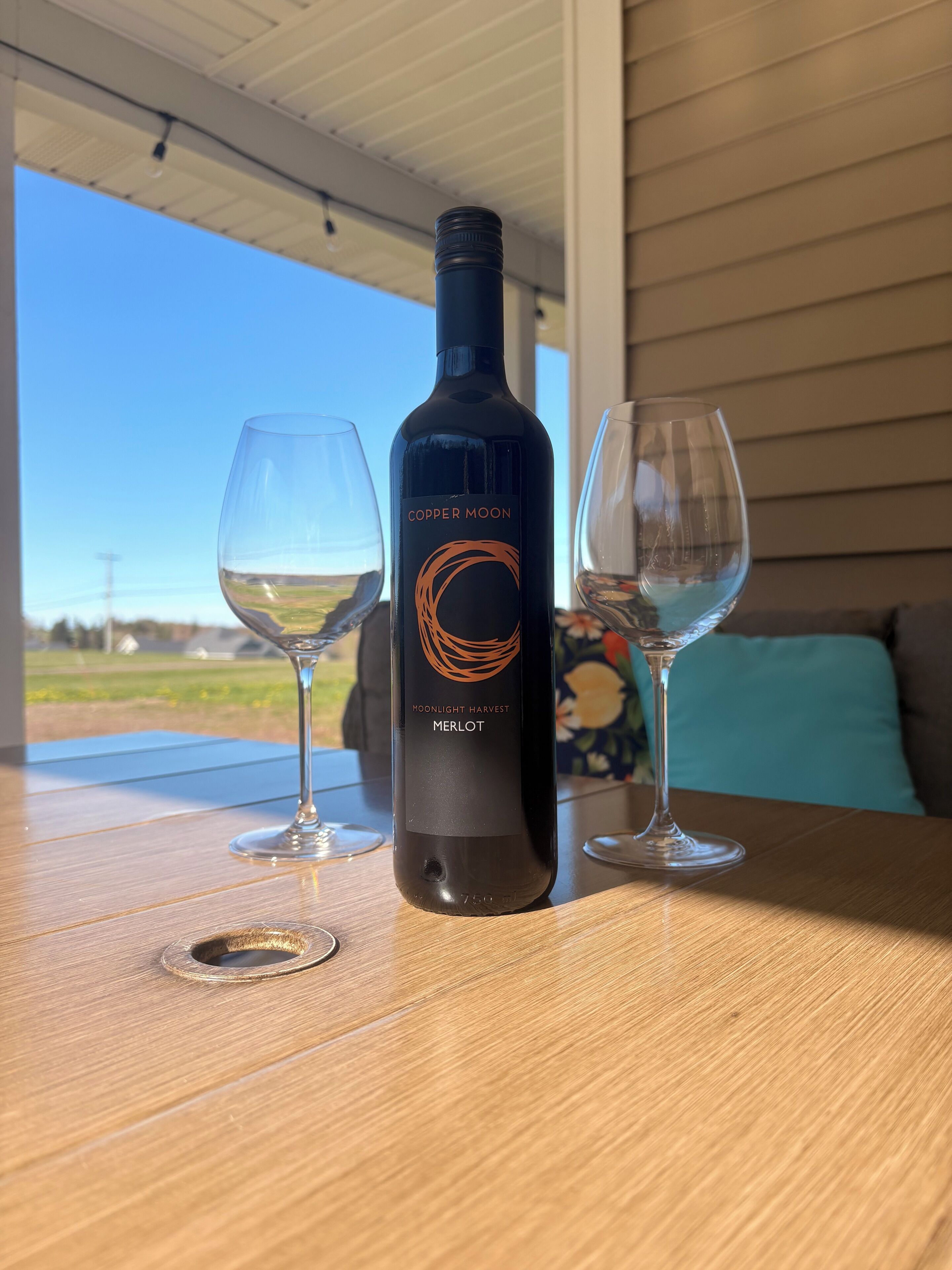 A bottle of Copper Moon Merlot wine on a table with two empty wine glasses.