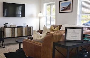 Un salon avec un canapé en cuir marron, un téléviseur à écran plat fixé au mur, une lampe et un certificat encadré posé sur une table.
