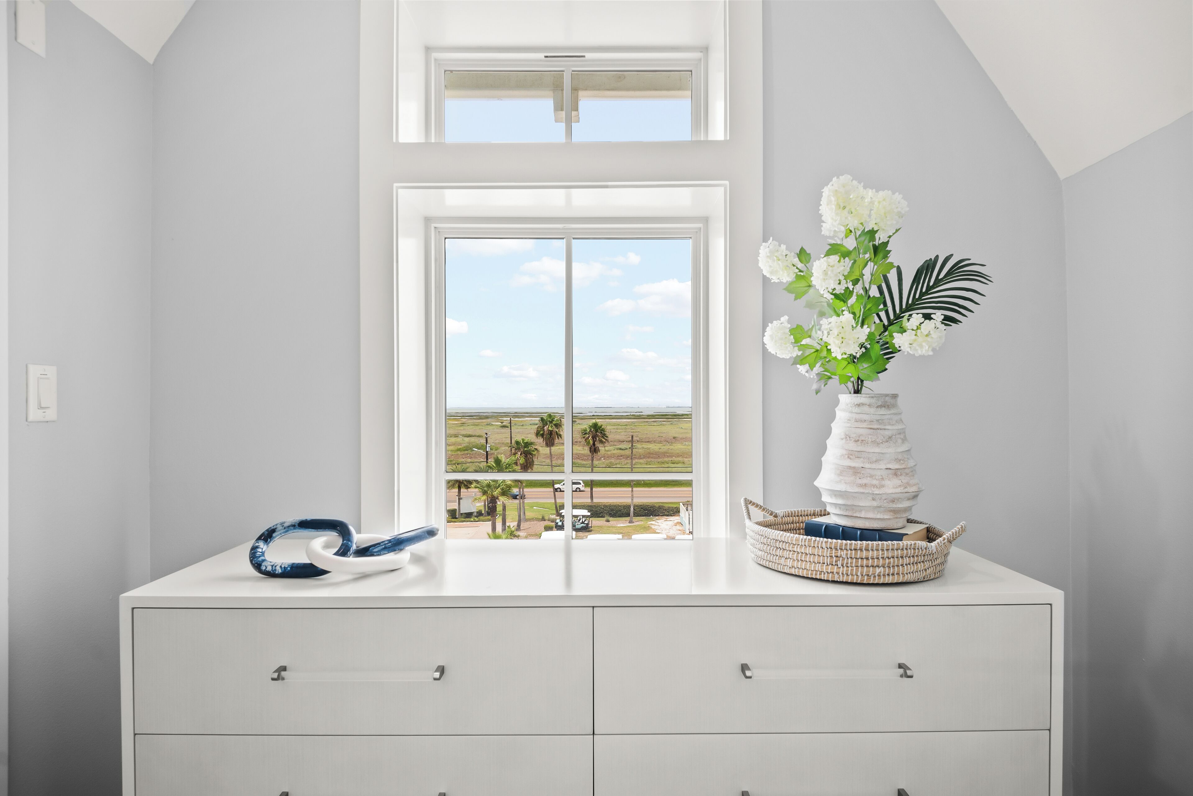 En vit byrå med lådor, en vas med vita blommor och ett palmblad, samt ett blått och vitt mönstrat föremål på byrån.