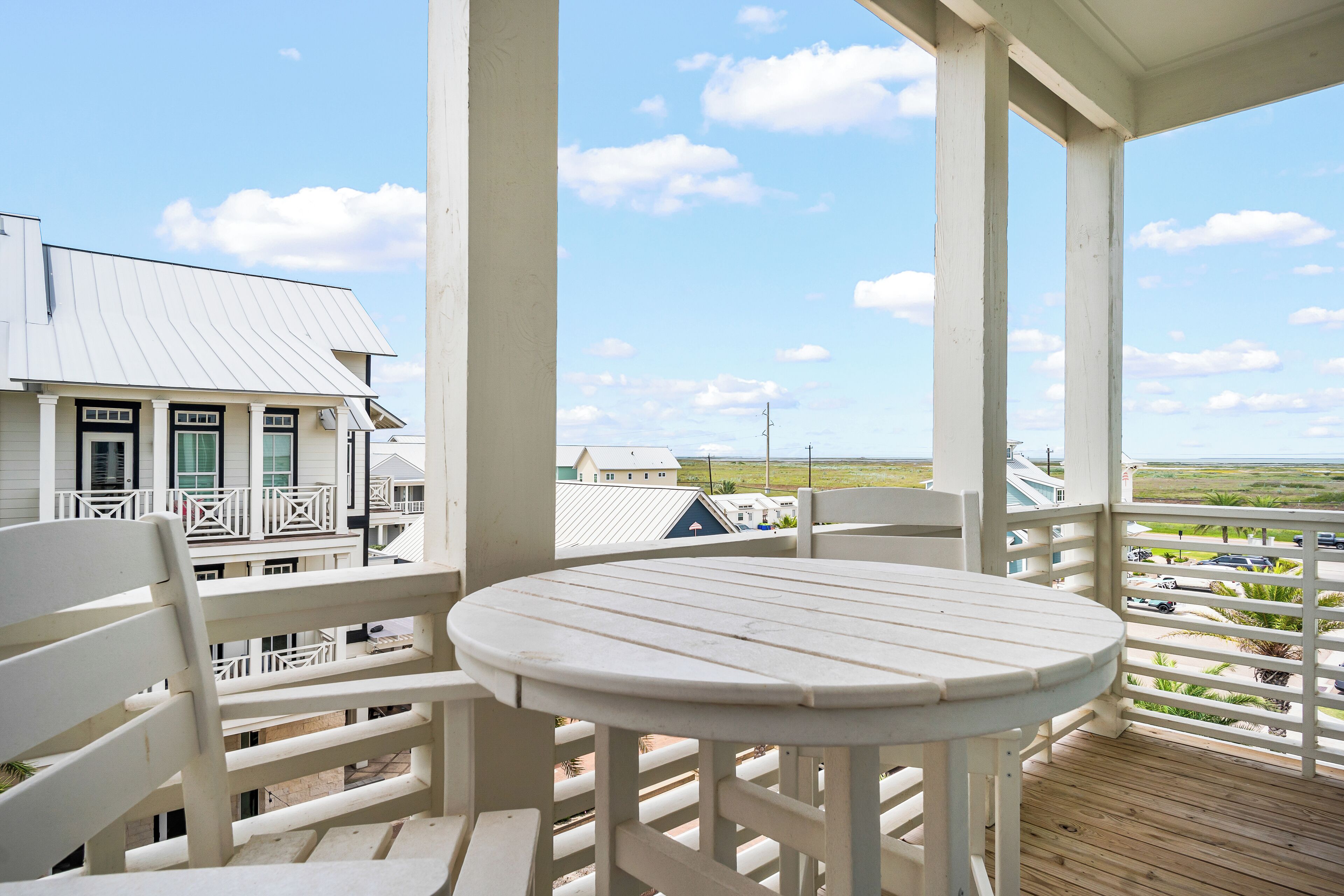 A covered outdoor area with a round table and white chairs, overlooking a residential area with houses and a clear sky.