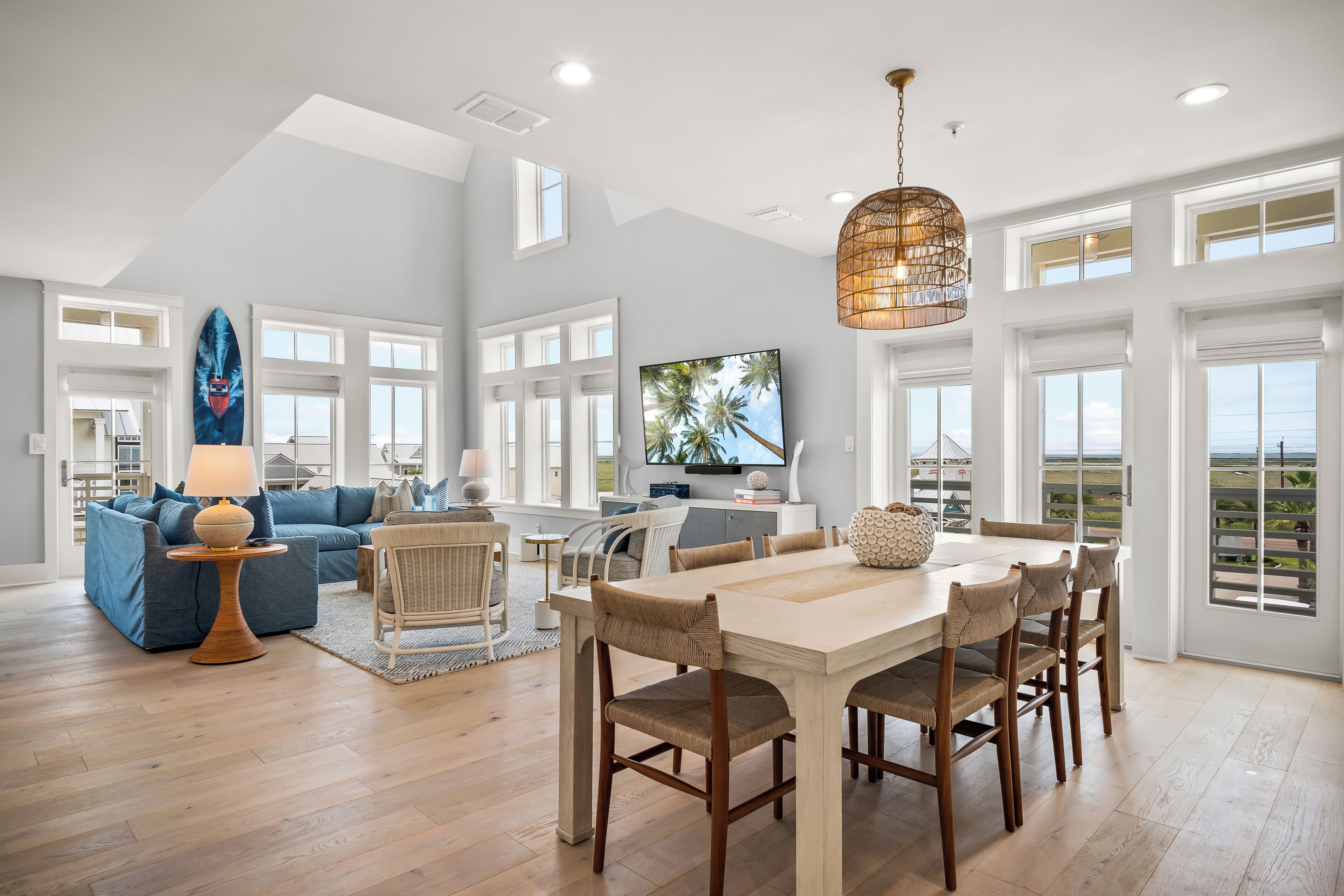 A spacious living room with a large dining table, comfortable seating, a television, and large windows offering a view of the outdoors.