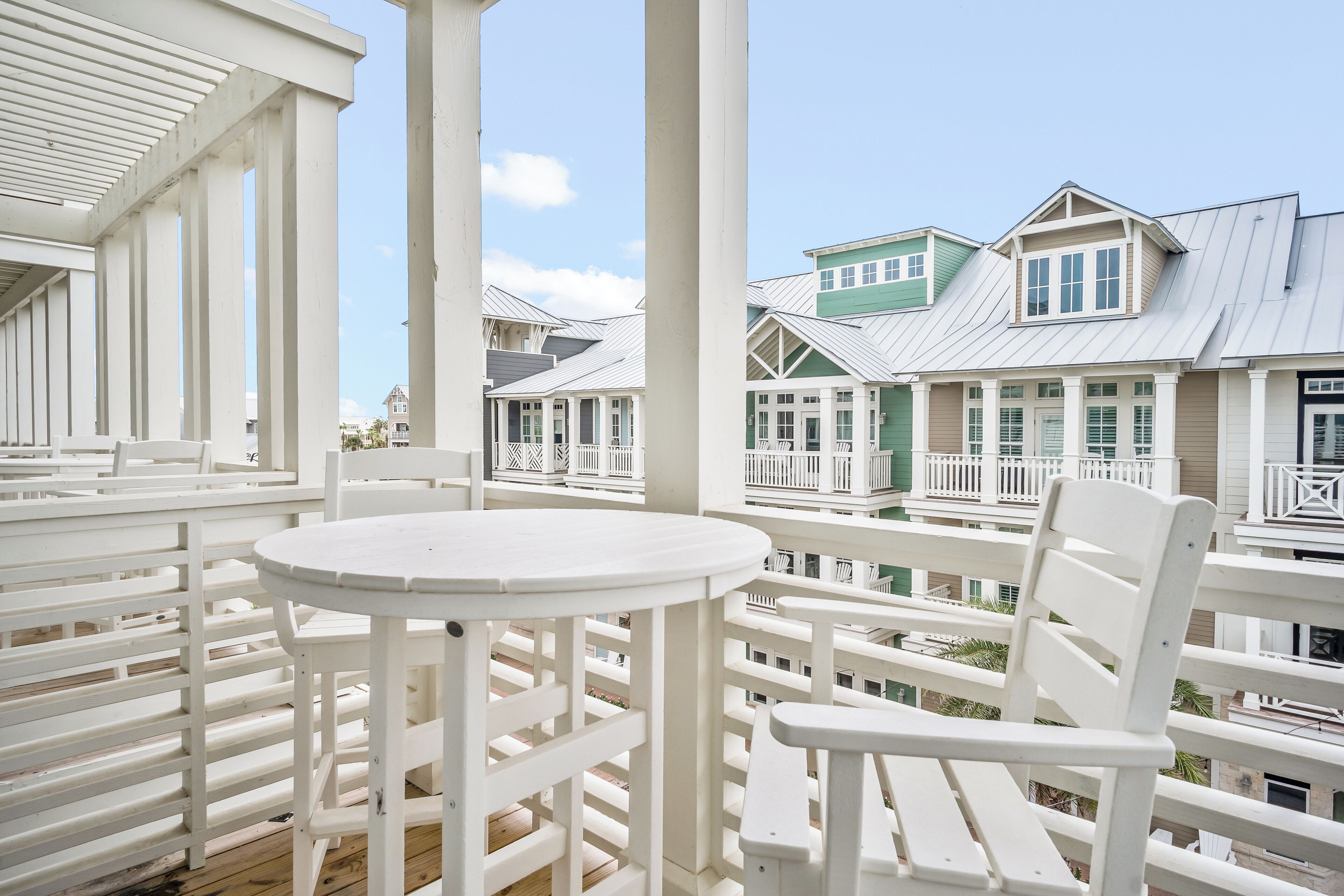 A balcony with white furniture, including a round table and chairs, overlooking a row of houses with white railings and green accents.