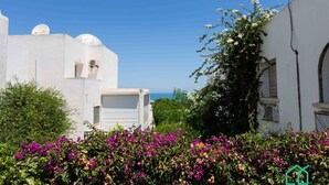 A white building with a satellite dish, a garage door, and a garden with purple flowers. - Le Duplex du Golden Tulip (La Marsa)