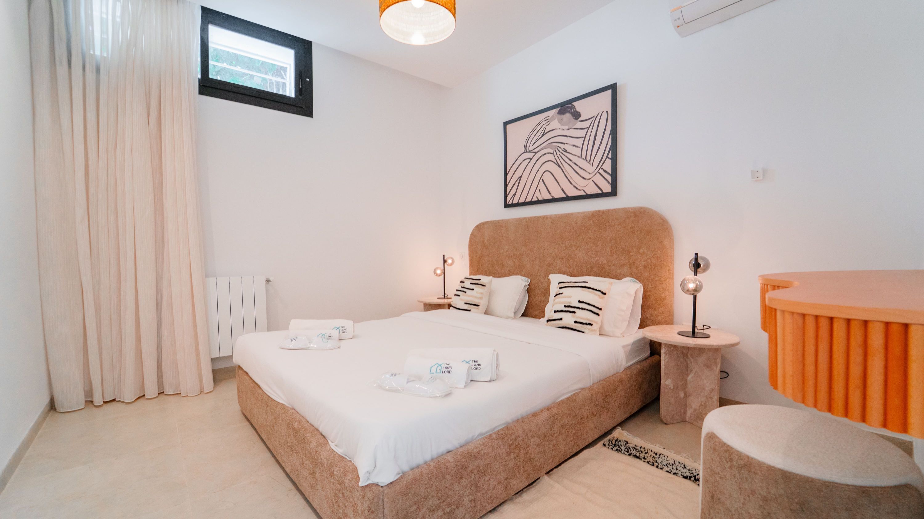 A bedroom with a large bed, a bedside table, a nightstand, a small round table, a mirror, a window, and a radiator.
