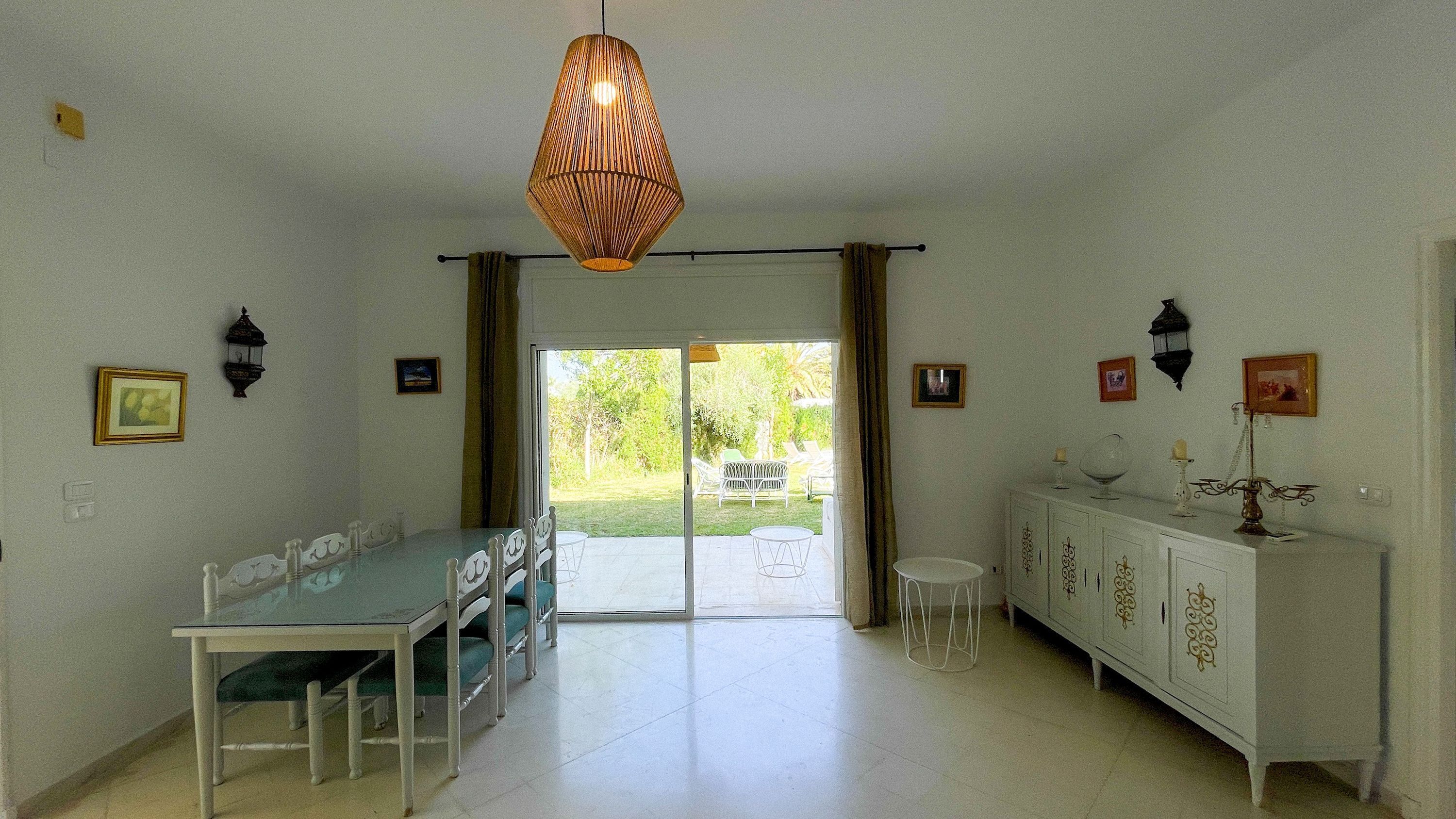 A dining area with a glass table and chairs, a white cabinet with decorative items, and a view of a garden through a sliding door.