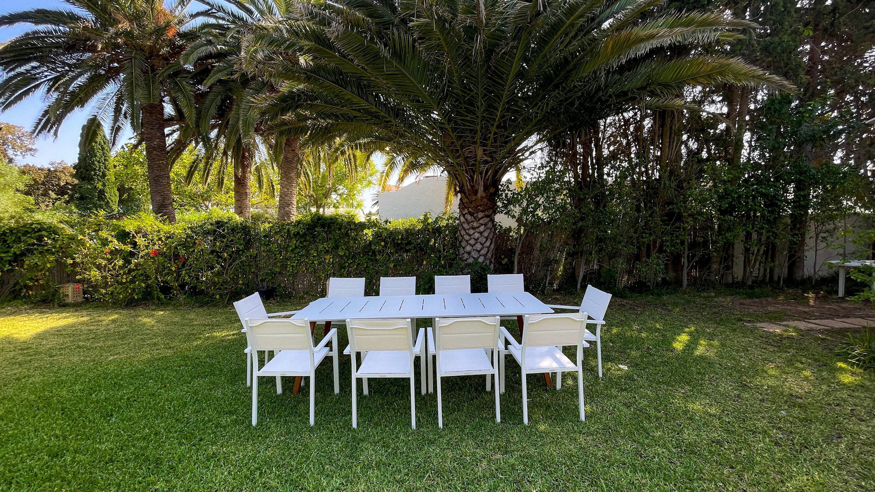 Uma área de refeições ao ar livre retangular, com mesa e cadeiras brancas, rodeada por vegetação exuberante e palmeiras.