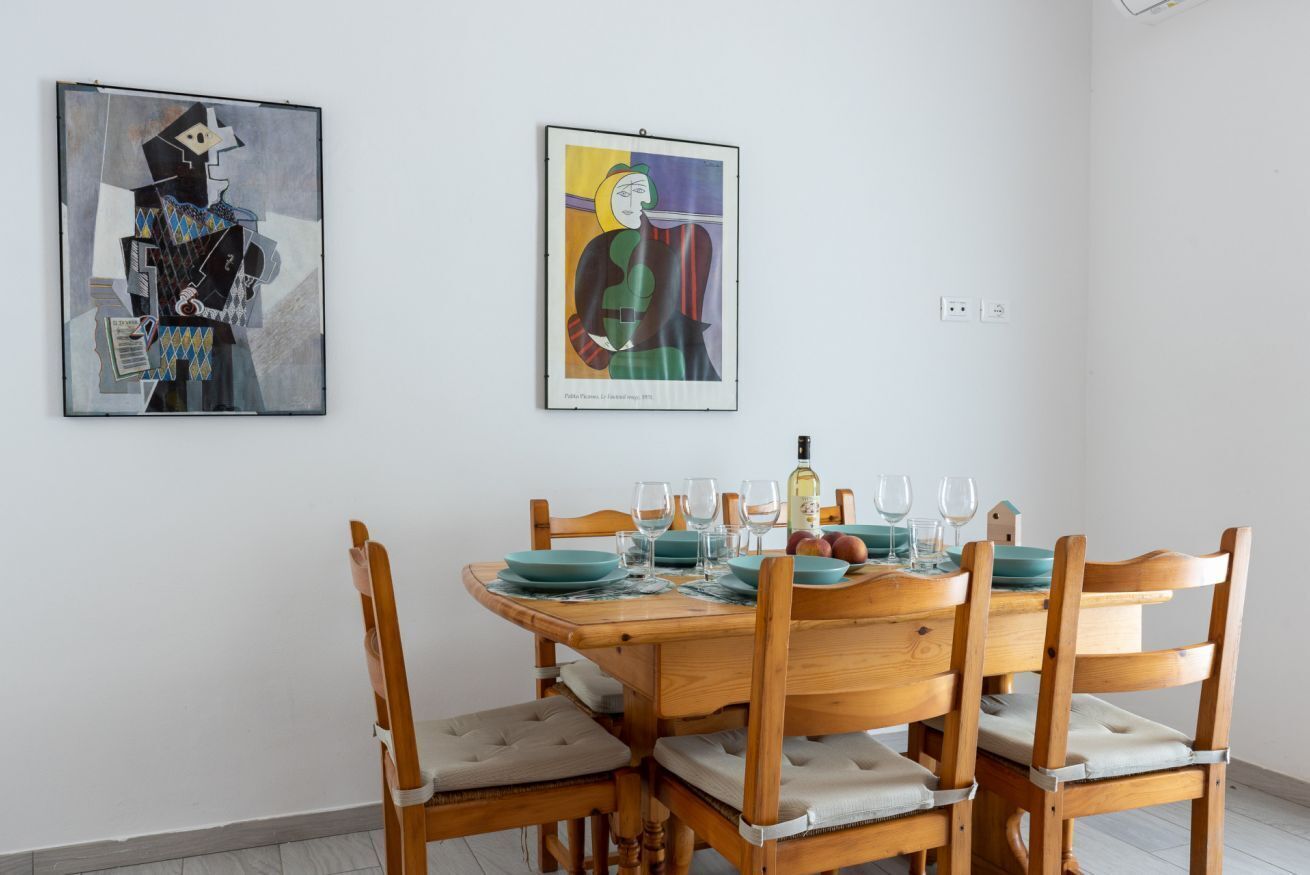 A dining area with a wooden table set for six, chairs with cushioned seats, and two framed artworks on the wall.