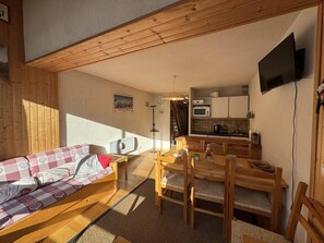 A cozy room with a wooden sofa, a dining table with chairs, a kitchen area, and a TV mounted on the wall.