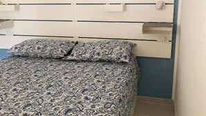 Ein Bett mit einem gemusterten Bettbezug und passenden Kissen, ein weißes Kopfende mit horizontalen Linien und ein kleines Regal mit einem weißen Gegenstand darauf.