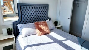 A neatly made bed with a tufted headboard, a fluffy throw pillow, and a patterned bedspread. There are bedside tables with small potted plants and a window providing natural light.