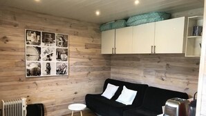 En kompakt opholdsstue med en sort sofa, et lille rundt bord og en radiator. En vægmonteret hylde rummer forskellige ting, og et hvidt skab er placeret over sofaen.