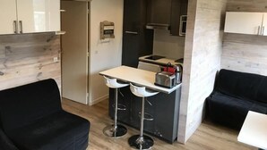 Et kompakt køkken med en hvid bordplade, to barstole og en sort sofa.