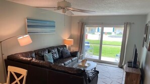 A living room with a dark leather sofa, a small table, and a ceiling fan. - Ocean View Ground Floor 2/2 (New Smyrna Beach)