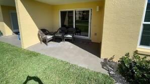 Un espacio al aire libre cubierto con sillas y una mesa, una puerta corrediza de vidrio y una pequeña zona ajardinada con césped y arbustos.