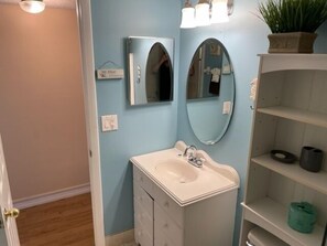 A bathroom with a white vanity, two wall-mounted mirrors, a light fixture above the vanity, and a shelf with a potted plant. - Ocean View Ground Floor 2/2 (New Smyrna Beach)