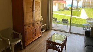 Una habitación con un armario de madera, una mesa con superficie de vidrio, un sofá y un ventanal corredero que da a un espacio exterior con sillas y césped.
