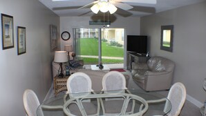 A living room with a glass-top table, wicker chairs, a beige sofa, a television, and a view of a green lawn through glass doors.