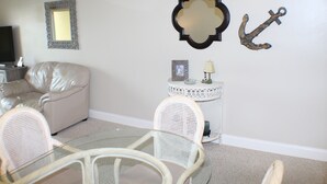 A living room with a beige sofa, a glass table, wicker chairs, a small side table with a lamp, and decorative wall art including a mirror and an anchor.
