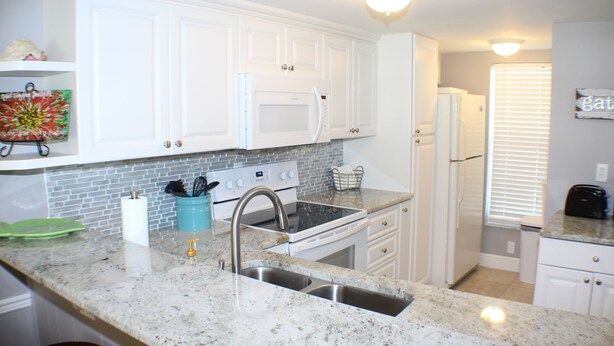 A kitchen with white cabinets, a granite countertop, a stainless steel sink, a microwave, an oven, and a refrigerator. - Ocean View Ground Floor 2/2 (New Smyrna Beach)