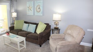 A living room with a brown sofa, a beige armchair, a small white table, and two wall-mounted lamps. - Ocean View Ground Floor 2/2 (New Smyrna Beach)