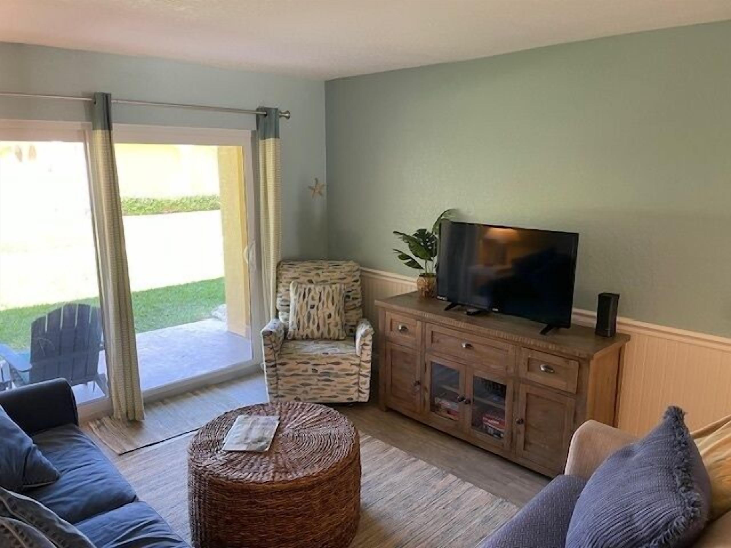 A living room with a sofa, a wicker ottoman, a television on a wooden cabinet, and a patterned armchair.