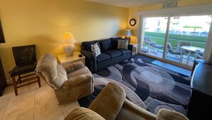 A living room with a sofa, armchairs, a coffee table, and a lamp. There is a sliding glass door leading to an outdoor patio area. - Ground Floor Ocean View 2/2 (New Smyrna Beach)