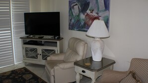 Een woonkamer met een televisie, een beige bank, een kamerkleed met een patroon en een schilderij aan de muur.