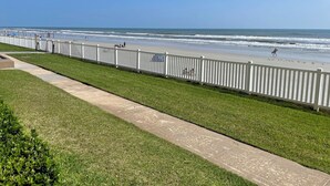 Une promenade en bord de mer, avec une clôture blanche, des zones herbeuses et une plage en arrière-plan.