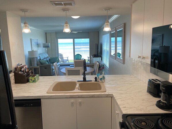 A kitchen with a double sink, a stove, and a coffee maker. There is a dining area with a table and chairs, and a view of the ocean through large windows.