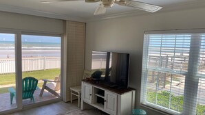 Ein Zimmer mit Deckenventilator, einem Fernseher auf einer Holzkabine, einer Schiebeglastür zum Balkon und Blick auf den Strand.