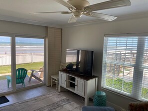 A room with a ceiling fan, a TV on a wooden cabinet, a sliding glass door to a balcony, and a view of the beach.