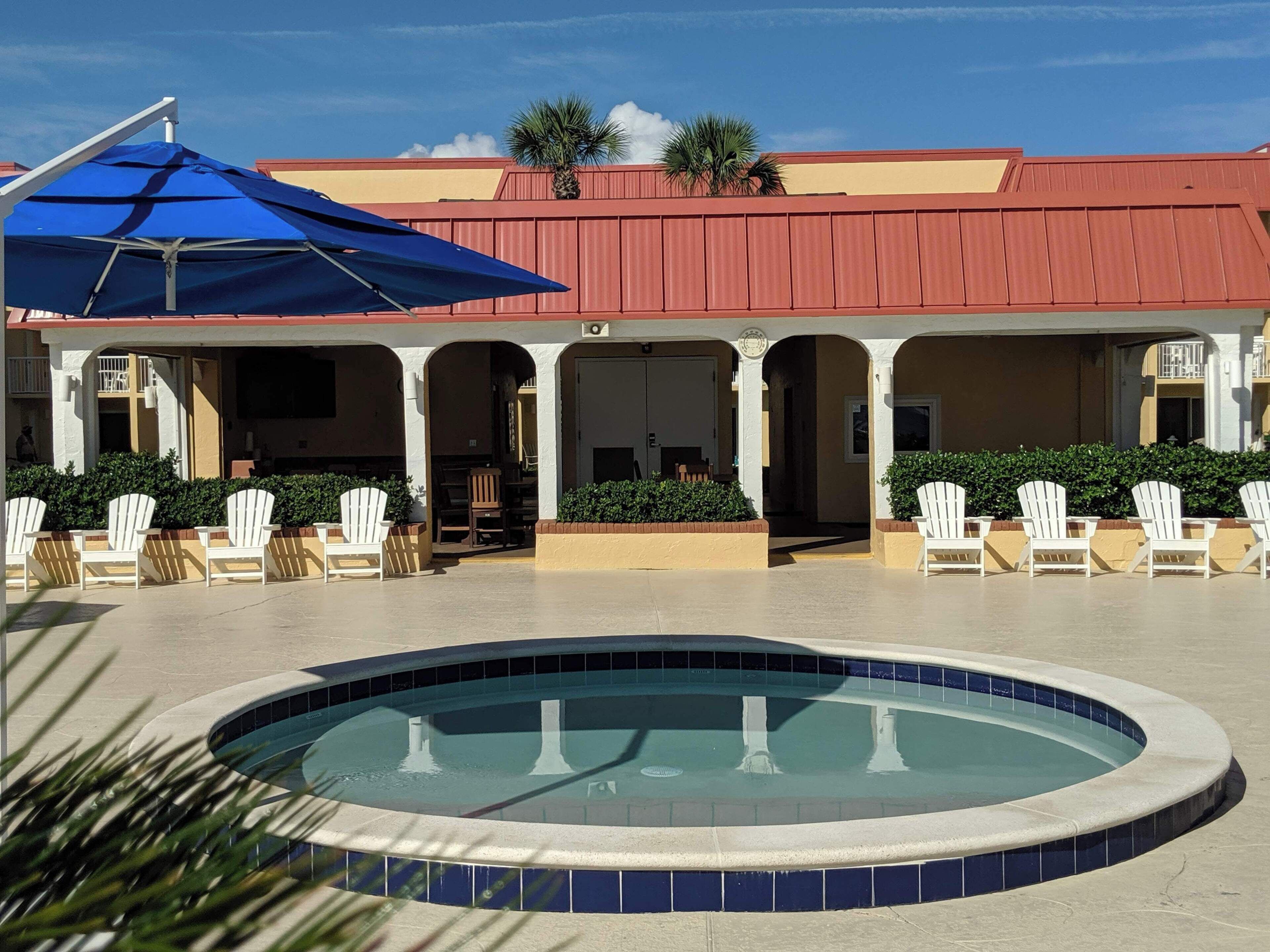 A pool area with a blue umbrella, white chairs, and a building with a red roof.