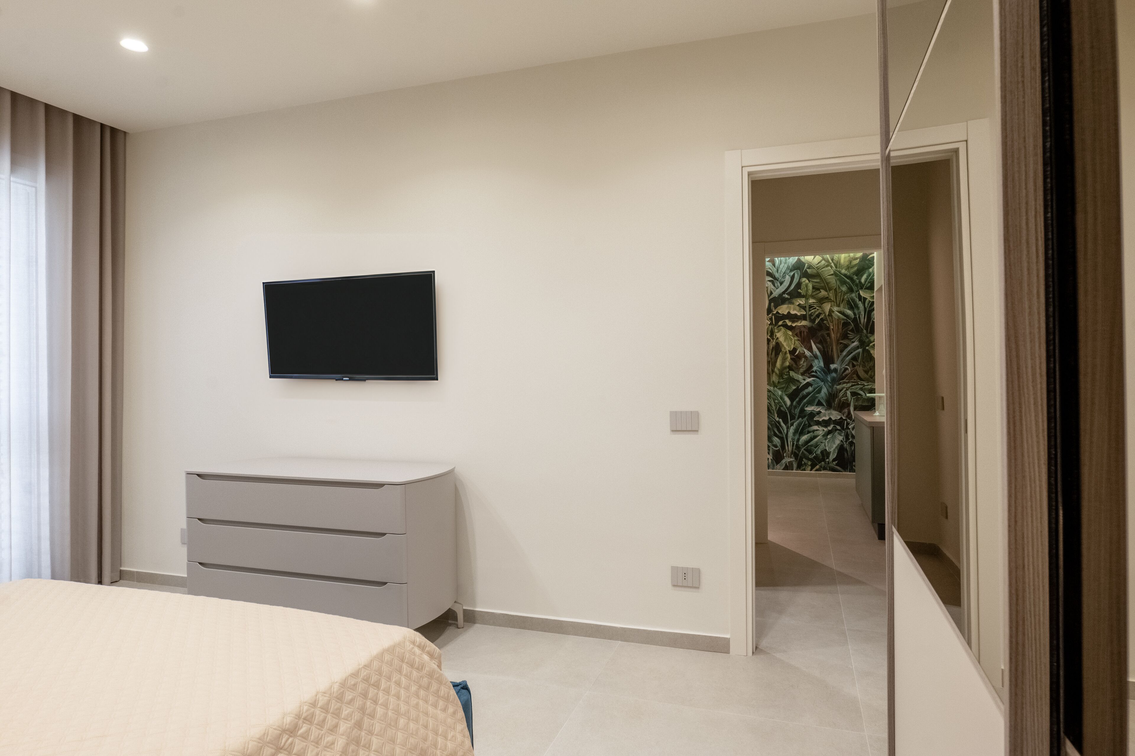 Une chambre à coucher comprenant un lit, une commode, un téléviseur à écran plat fixé au mur et une porte donnant sur un balcon agrémenté de plantes.