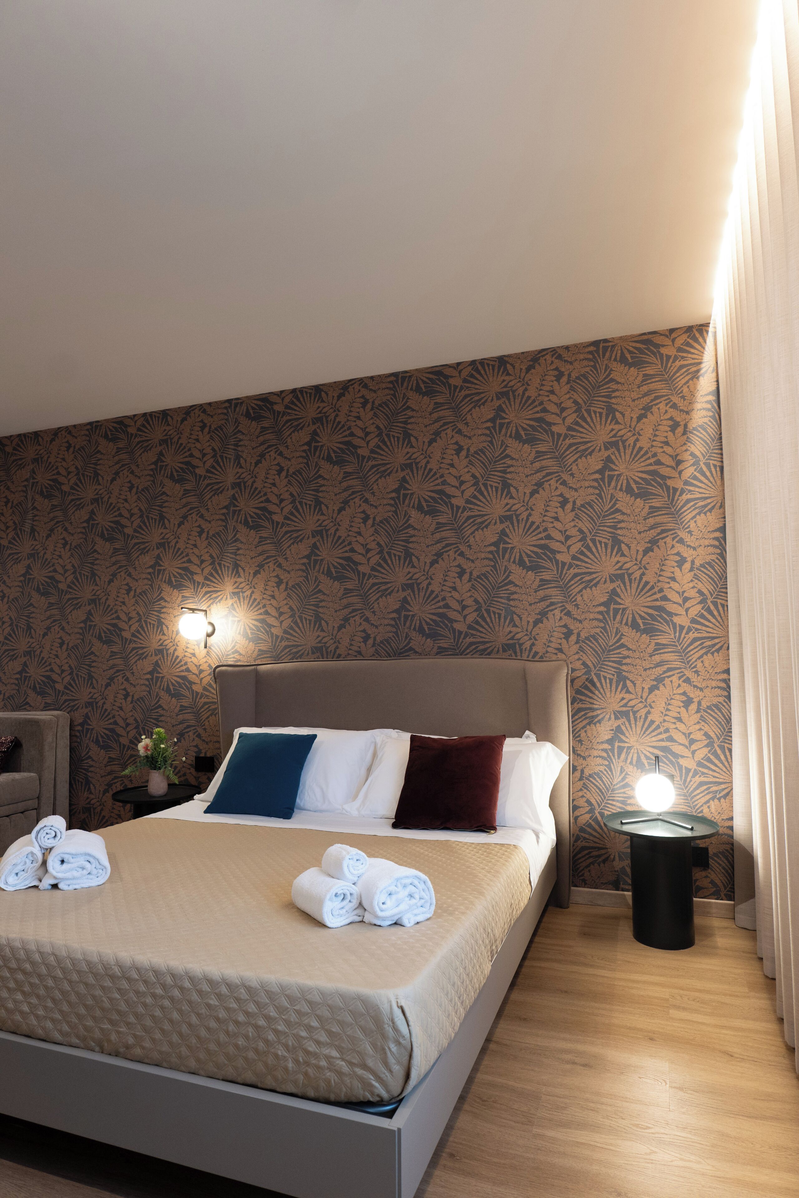 A neatly made bed with rolled towels, a bedside table with a lamp, and a patterned wallpaper.