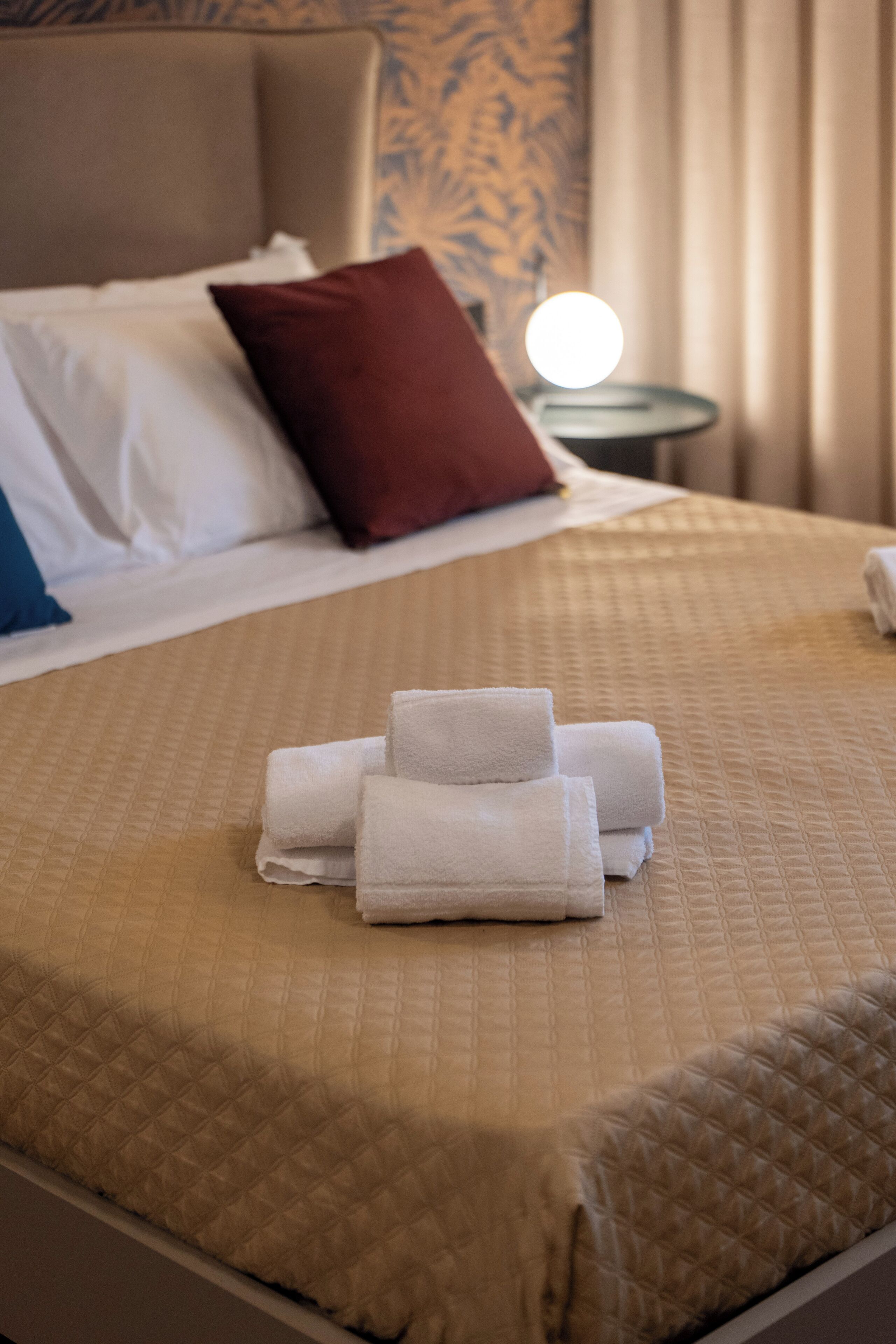 A neatly made bed with a beige quilt, a red pillow, and a stack of white towels on the bed.