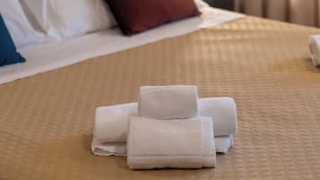 A neatly made bed with a beige quilt, a red pillow, and a stack of white towels on the bed.