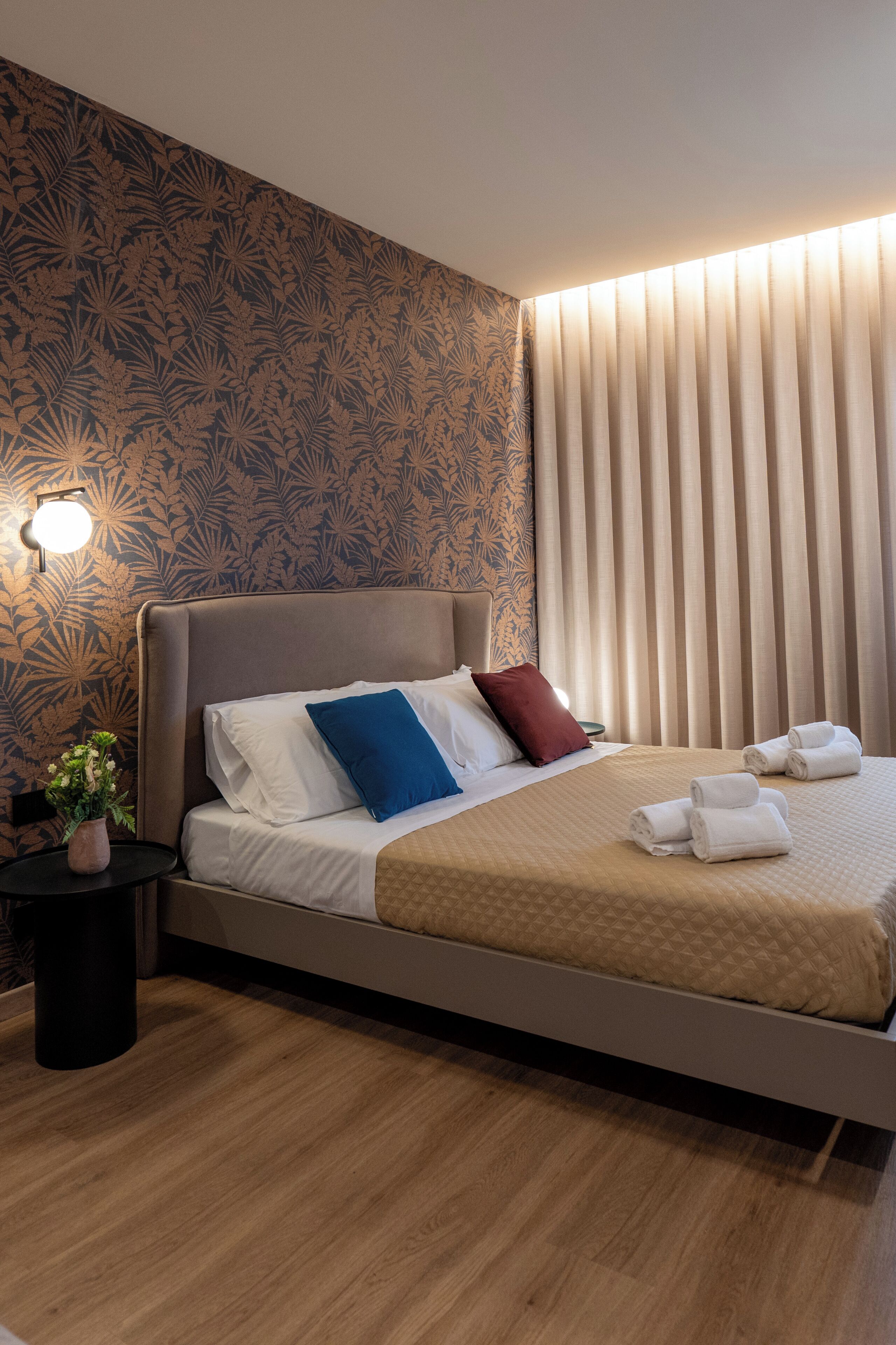 A modern hotel room with a large bed, a nightstand with a vase of flowers, and a window with vertical blinds.