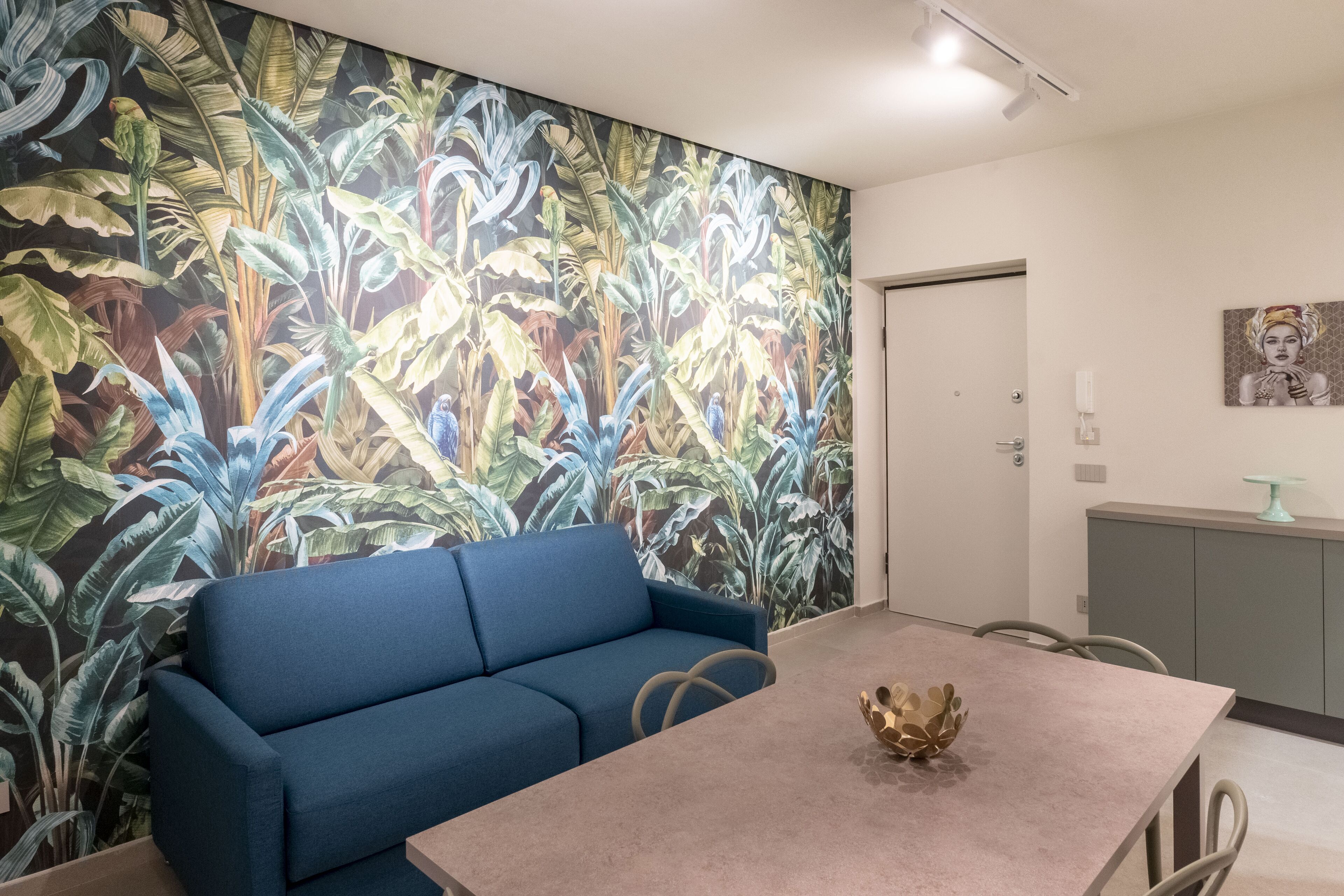 Una habitación con un sofá azul, una mesa con un cuenco decorativo, un mural con plantas tropicales y un armario gris con cubierta de vidrio.