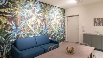 Una habitación con un sofá azul, una mesa con un cuenco decorativo, un mural con plantas tropicales y un armario gris con cubierta de vidrio.