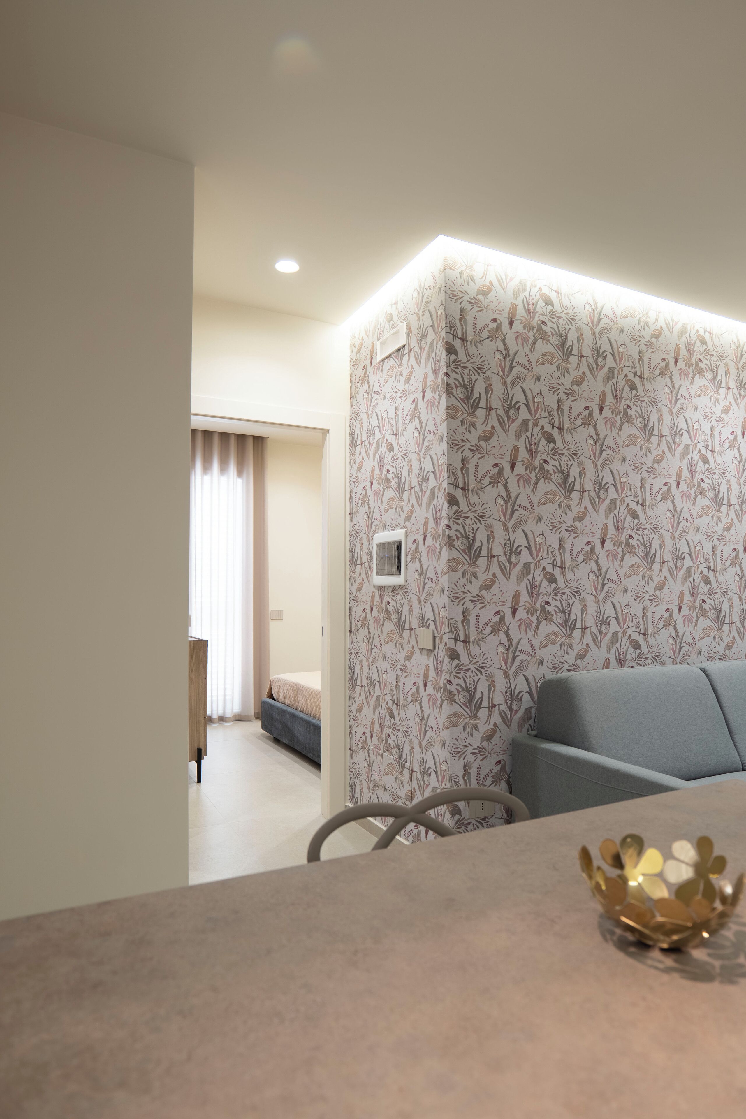 Ein modernes Hotelzimmer mit einer grauen Couch, einem kleinen Tisch mit einer dekorativen Schale und einer gemusterten Tapete.