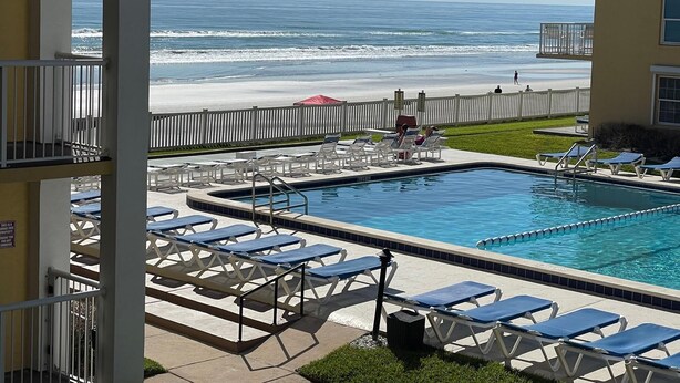 A pool area with lounge chairs, a view of the ocean, and a fenced beach.