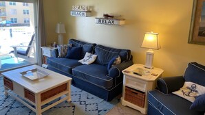 A living room with a sofa, a coffee table, a side table, and a lamp. There are two signs on the wall, one reads 'ESCAPE BEACH' and the other 'RELAX'.