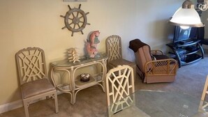 A room with a glass-top table, two wicker chairs, a decorative ship's wheel, and a table lamp. - Ocean View Second Floor 2/2 (New Smyrna Beach)