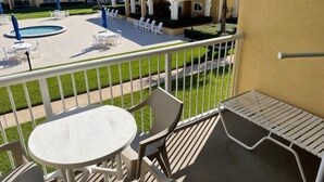 A balcony with a round table and chairs, a pool area, and a building with a red roof in the background. - Ocean View Second Floor 2/2 (New Smyrna Beach)