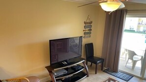 A living room with a flat-screen TV on a wooden stand, a glass coffee table, a wicker sofa, a black chair, and a ceiling fan. - Ocean View Second Floor 2/2 (New Smyrna Beach)