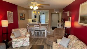 A living room with floral-patterned sofas, a dining table with chairs, a ceiling fan, and a kitchen area in the background. - Ocean View  Second Floor 2/2 (New Smyrna Beach)