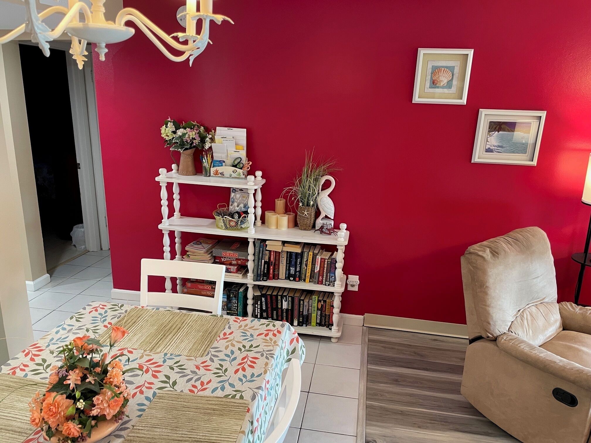 Une pièce avec un mur rouge, une bibliothèque blanche, un fauteuil beige et une table recouverte d’une nappe à motifs floraux.