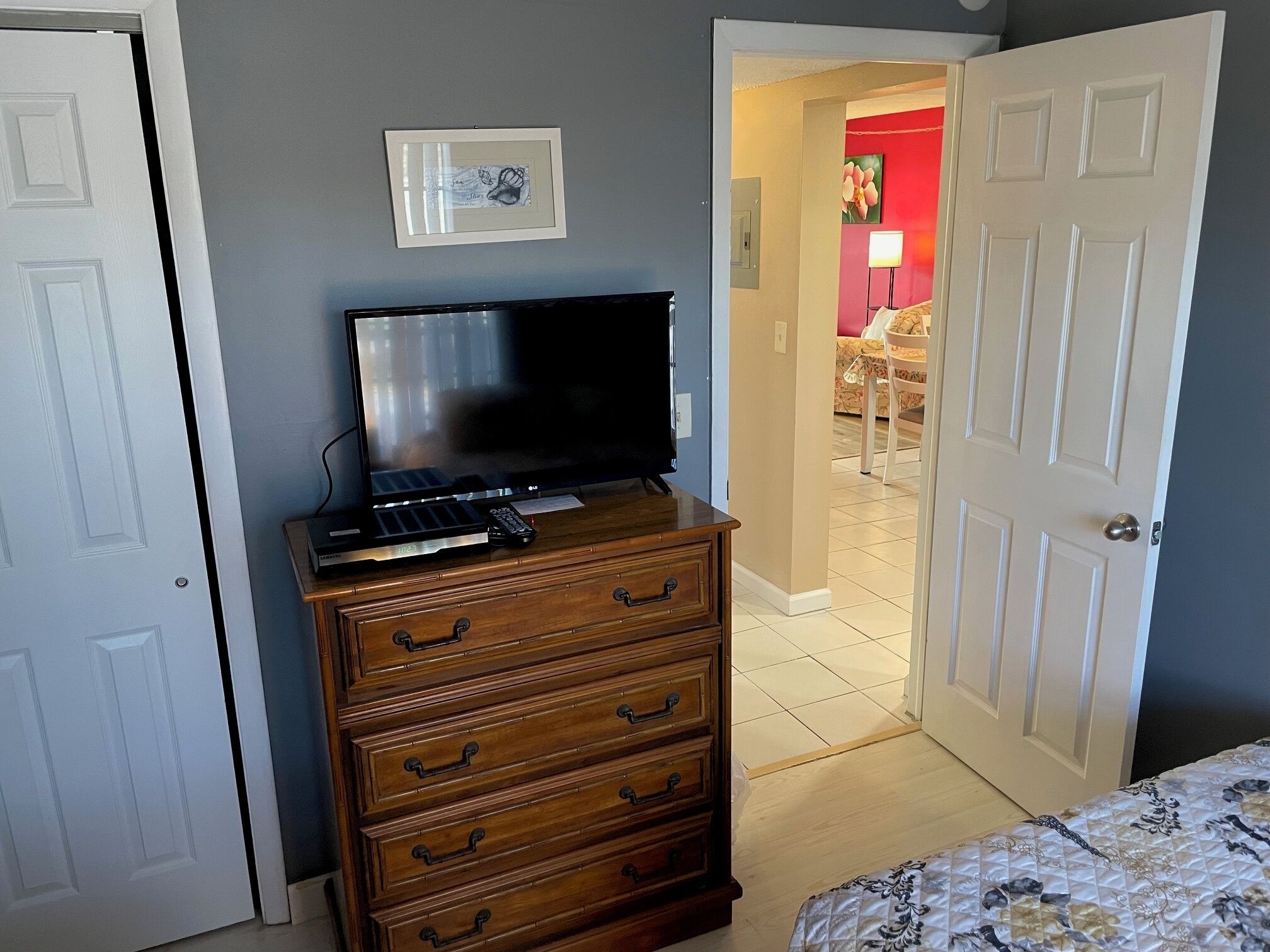 Een slaapkamer met een televisie op een houten dressoir, een bed met een bloemenbedekking, en een deur die leidt naar een zithoek.